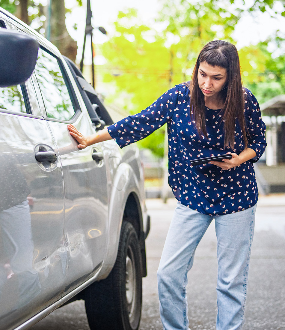 A woman holding a tablet while inspecting the damage on her car