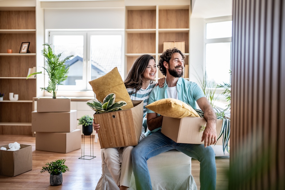 A happy young couple holding boxes with plants and pillows as they move into their new home