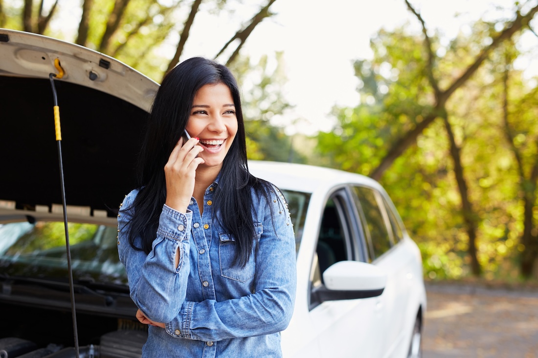 A smiling woman calls a repair shop on her phone as she stands beside a car with its hood up.