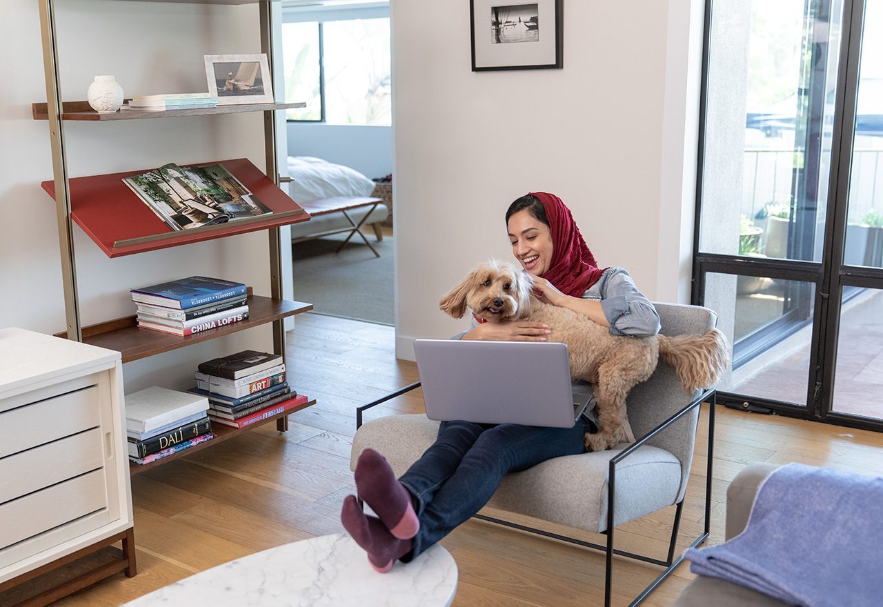 A woman sits with a laptop and her dog on her lap in the living room of her home. disaster planning pets