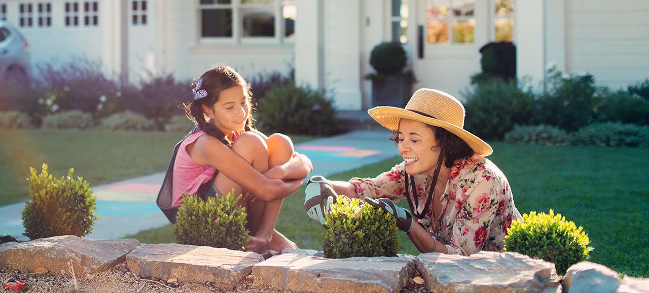 A woman trims a small shrub in her front yard with garden clippers while her granddaughter watches. yard maintenance