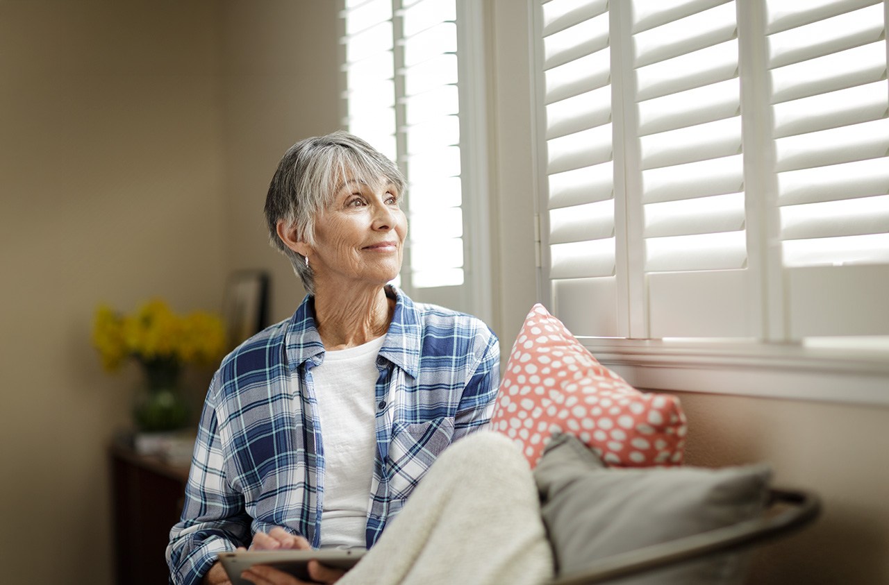 A woman in a blue flannel shirt sits on a window seat looking out the window of her home. renters insurance
